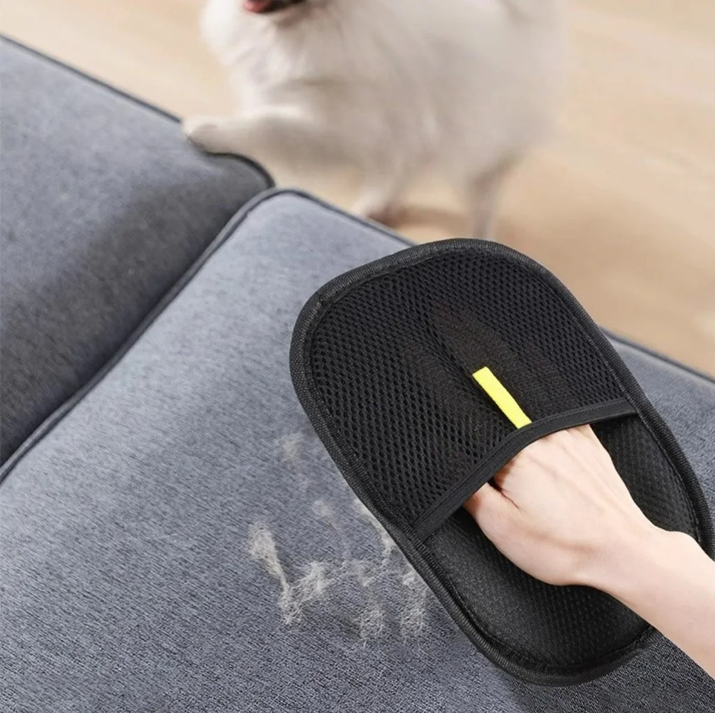 Person using a black pet grooming brush on a gray couch with a dog in the background.