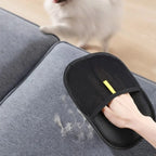 Person using a black pet grooming brush on a gray couch with a dog in the background.
